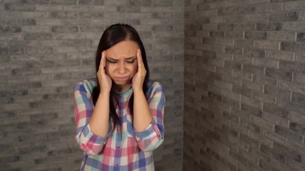 Een jonge vrouw in een geruit shirt grijpt haar hoofd met handen vol pijn op een bakstenen achtergrond. Een vrouw heeft hoofdpijn.. — Stockvideo
