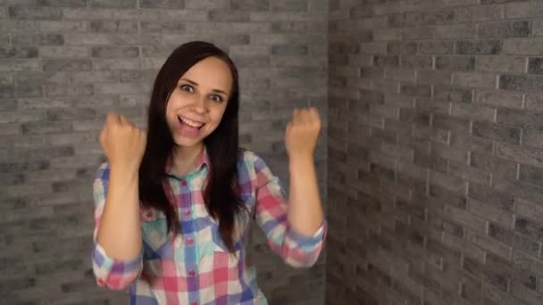 Una guapa joven en una camisa de camisa de camisa se ríe alegremente y expresa emociones positivas agitando sus puños sobre un fondo de ladrillo gris.. — Vídeo de stock