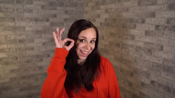 Uma jovem mulher bonita em um vermelho hoodie mostra está sorrindo e mostrando sinal de OK em um fundo de tijolo cinza. — Vídeo de Stock