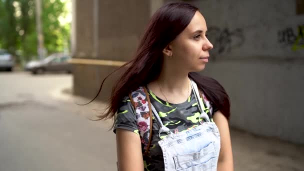 Una guapa joven vestida con ropa casual sonríe en la calle. La larga morena se desarrolla en el viento.. — Vídeos de Stock