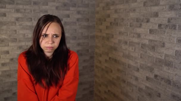 A beautiful young woman in a red hoodie is being in bad mood. A young woman shows a sadness on a brick background. – Stock-video