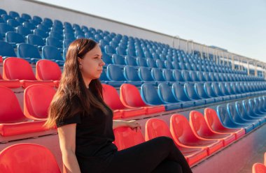 Uzun saçlı, siyah elbiseli genç bir kadın stadyum tribünlerinde tek başına oturuyor ve kavurucu güneşin altında spor maçı izliyor..