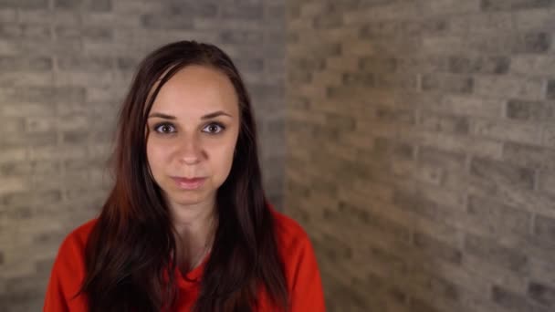 A young woman in a red hoodie shows with insult her tongue looking at camera on a brick background. – Stock-video