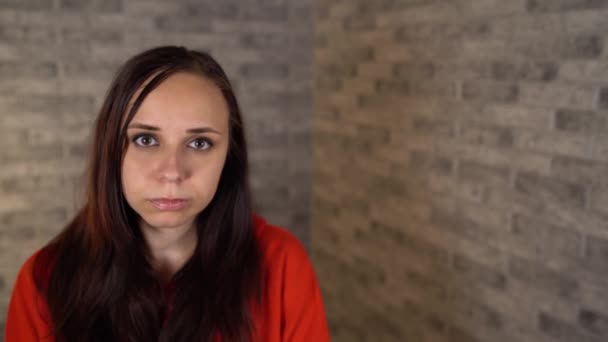 En vacker ung kvinna i röd luvtröja är på dåligt humör. En ung kvinna visar en sorg på en tegelsten bakgrund. — Stockvideo