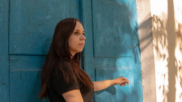 Uma mulher jovem em um t-short preto com estandes longos do cabelo e olha na distância no fundo de um edifício velho com portas azuis. — Fotografia de Stock