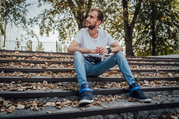 Un joven guapo con ropa casual está sentado en los escalones del parque y mirando a su alrededor.. — Foto de Stock