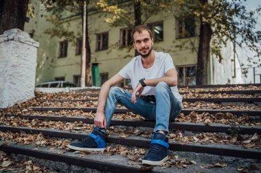 Gündelik giysiler içinde genç yakışıklı bir adam parkta merdivenlerde oturuyor ve kameraya bakıyor. Metin için alanı kopyala.
