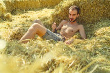 Sakallı genç yakışıklı bir adam samanlıkta çıplak bir gövdeyle yatıyor, elinde bir kemer tutuyor ve kameraya flört ederek bakıyor..