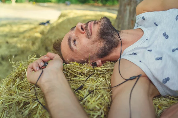 Un giovane uomo bello con barba in t-shirt e pantaloncini giace nel fieno, sorridendo e ascoltando musica — Foto Stock