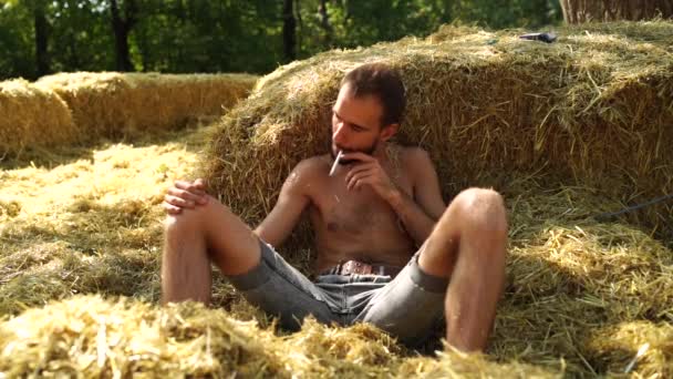 髭を生やした若いハンサムな男が裸の胴で干し草の中に横たわってタバコを吸っている — ストック動画