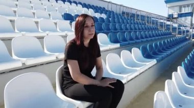 Siyah elbiseli, uzun saçlı genç bir kadın stadyumda oturuyor.