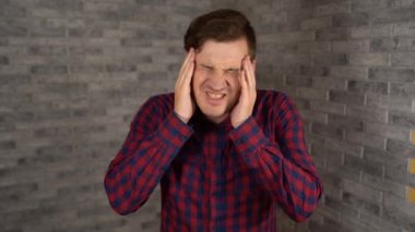 Ekose gömlekli genç ve güzel bir adam gri tuğlalı arka planda acı içinde ellerini şakaklarına koyuyor..