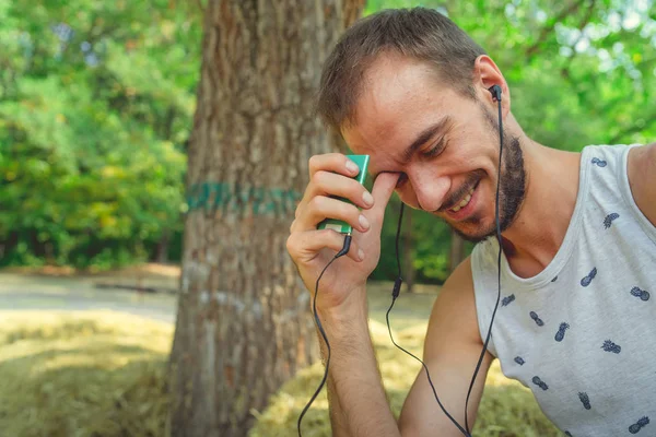 Un giovane uomo bello con barba in t-shirt si siede nel fieno, ascolta musica e canta nel parco. — Foto Stock