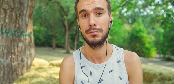 Un joven guapo con barba en una camiseta se sienta en el heno, escucha música y canta en el parque.. — Foto de Stock