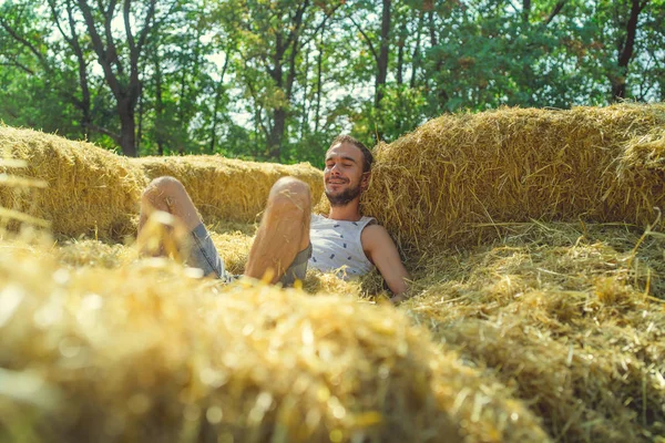 Un joven guapo con barba de delgada construcción en una camiseta y pantalones cortos se encuentra en el heno en un día soleado.. — Foto de Stock