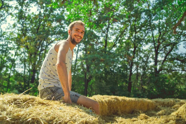 Un joven y guapo hombre con barba de construcción delgada se sienta en el heno y sostiene el heno en sus manos sobre el fondo de los árboles verdes del parque.. — Foto de Stock