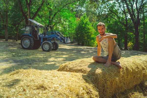 Um homem bonito novo com barba da construção slim senta-se no feno e prende o feno em suas mãos de encontro ao fundo das árvores verdes no parque. — Fotografia de Stock