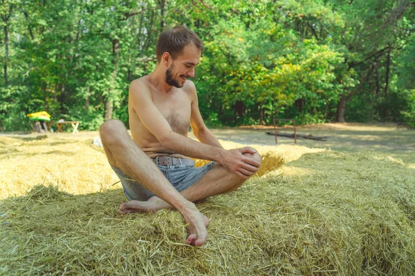 Un joven guapo con barba y un torso desnudo se sienta en el heno y sonríe sobre el fondo de los árboles verdes del parque.. — Foto de Stock