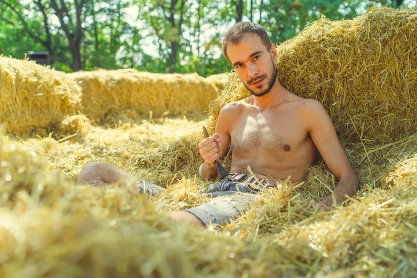 Un joven guapo con barba y un torso desnudo se sienta en el heno y sonríe sobre el fondo de los árboles verdes del parque.. — Foto de Stock