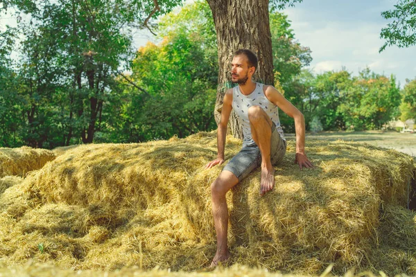 Un joven y guapo hombre con barba de construcción delgada se sienta en el heno y sostiene el heno en sus manos sobre el fondo de los árboles verdes del parque.. — Foto de Stock