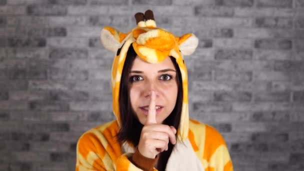 Una jovencita divertida en un gran pajamas de jirafa sonríe y hace gesto silencioso con el dedo índice en los labios sobre un fondo de ladrillo gris. El concepto de silencio. — Vídeo de stock