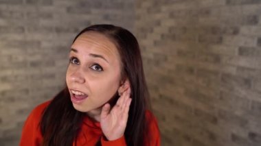 Kırmızı kapüşonlu genç bir kadın kulağına el ele tutuşuyor ve gri tuğlalı bir arka planda konuşmaya kulak misafiri olmaya çalışıyor. Meraklı bir esmer geniş bir şekilde gülümsüyor ve birini gözetliyor. Konsept: