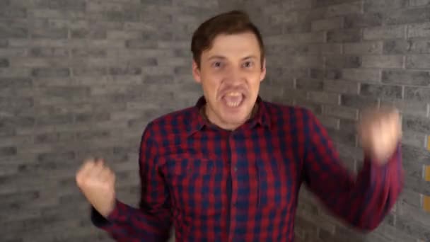 격자무늬 셔츠를 입은 젊고 잘생긴 남자가 행복하게 웃으면서 회색 벽돌 배경에 주먹을 흔들며 긍정적 인 감정을 표현하고 있다. — 비디오