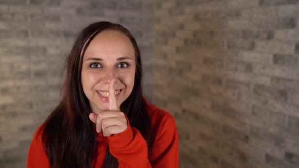 En ung kvinna i röd huvtröja håller på att tysta gester med pekfingret mot läpparna på en grå tegelsten bakgrund. Begreppet tystnad. — Stockvideo