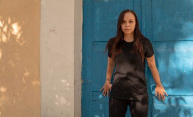 Siyah tişörtlü, uzun saçlı, mavi kapılı, eski bir binanın arka planında genç bir kadın..