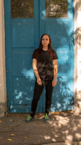 Siyah tişörtlü, uzun saçlı, mavi kapılı, eski bir binanın arka planında genç bir kadın.. — Stok fotoğraf
