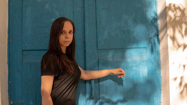 Uma jovem mulher em um t-short preto com cabelos longos no fundo de um edifício velho com portas azuis. — Fotografia de Stock