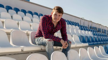 Ekose gömlekli ve kot pantolonlu genç yakışıklı bir adam stadyum tribünlerinde tek başına oturuyor, gülümsüyor ve yazın başka tarafa bakıyor..