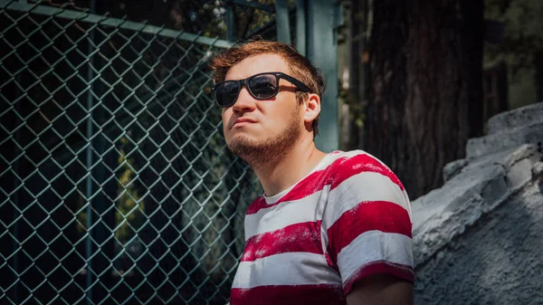 A portrait of a young handsome man in a t-shirt and in sunglasses near a lattice fence in the park. — Stock Photo, Image