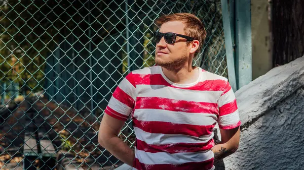 Un retrato de un joven guapo en una camiseta y en gafas de sol cerca de una valla de lattice en el parque.. — Foto de Stock