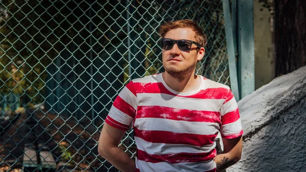 Un retrato de un joven guapo en una camiseta y en gafas de sol cerca de una valla de lattice en el parque.. — Foto de Stock