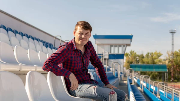 Een jonge knappe man in geruit hemd en jeans zit alleen op een tribune, glimlacht en kijkt weg in de zomer. — Stockfoto