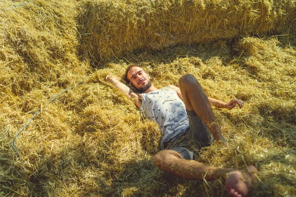 Un joven guapo con barba de delgada construcción en una camiseta y pantalones cortos se encuentra en el heno en un día soleado.. — Foto de Stock