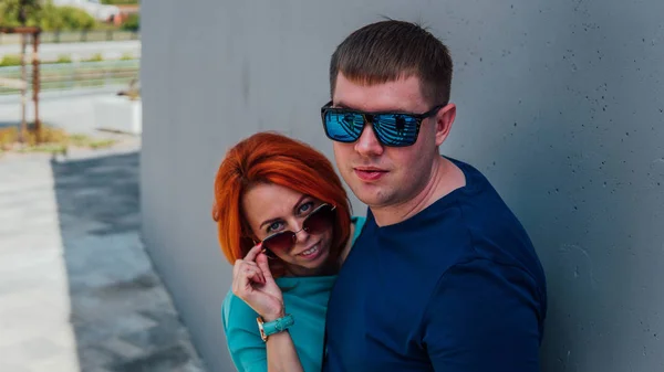 Ευτυχισμένο ζευγάρι με καθημερινά ρούχα είναι σε μια βόλτα. Ευτυχισμένος άντρας και γυναίκα. Ένας άντρας με γυαλιά ηλίου και μια κοκκινομάλλα στον πέτρινο τοίχο.. — Φωτογραφία Αρχείου