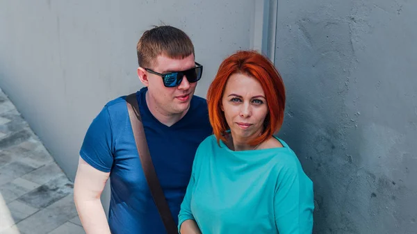 Feliz pareja vestida con ropa ocasional están de paseo. Feliz hombre y mujer. Un hombre en gafas de sol con mujer de pelo rojo contra una pared de piedra.. — Foto de Stock