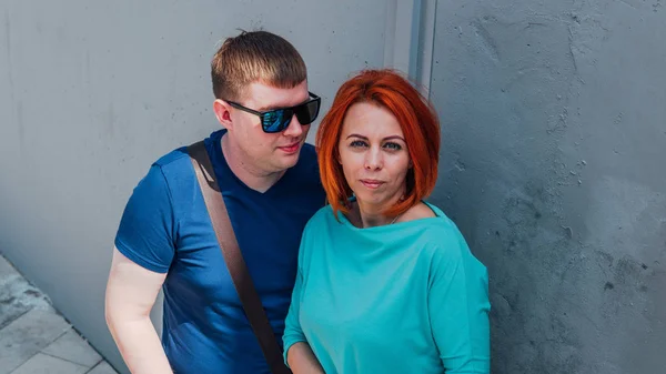 Feliz pareja vestida con ropa ocasional están de paseo. Feliz hombre y mujer. Un hombre en gafas de sol con mujer de pelo rojo contra una pared de piedra.. — Foto de Stock