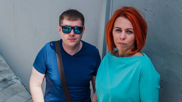 Feliz pareja vestida con ropa ocasional están de paseo. Feliz hombre y mujer. Un hombre en gafas de sol con mujer de pelo rojo contra una pared de piedra.. —  Fotos de Stock