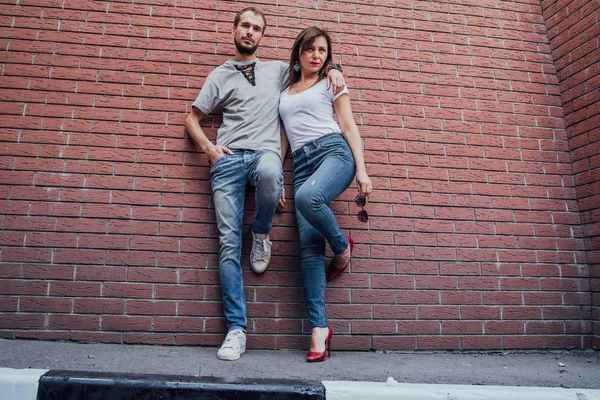 Un jeune couple amoureux en t-shirts et en jeans pose contre un mur de brique. — Photo