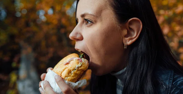 O femeie mănâncă un burger într-un parc însorit de primăvară. Tânără femeie cu soare zi mănâncă hamburger în pădurea de toamnă . — Fotografie, imagine de stoc