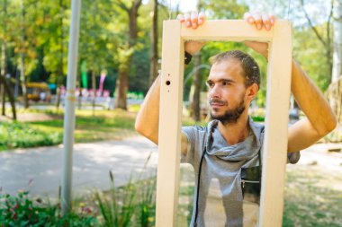 Parkta sıradan giysiler içinde sakallı yakışıklı bir adamın portresi. Adam asılı bir pencerenin arkasında durup kameraya poz veriyor..