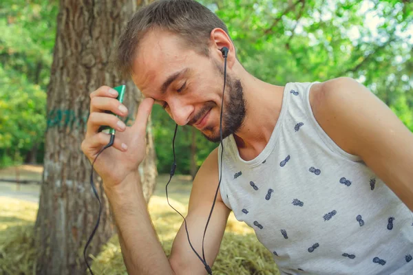 Un giovane uomo bello con barba in t-shirt si siede nel fieno, ascolta musica e canta nel parco. — Foto Stock