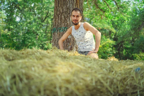 Un joven y guapo hombre con barba de construcción delgada se sienta en el heno y sostiene el heno en sus manos sobre el fondo de los árboles verdes del parque.. — Foto de Stock