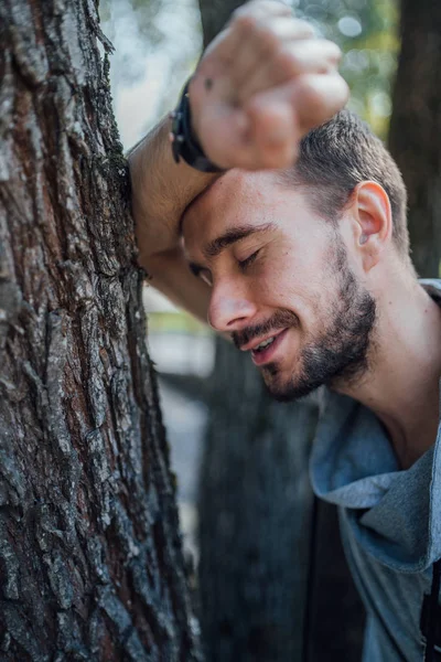 Un retrato de un hombre guapo con barba recostando su brazo contra un árbol con ojos cerrados en el parque.. — Foto de Stock