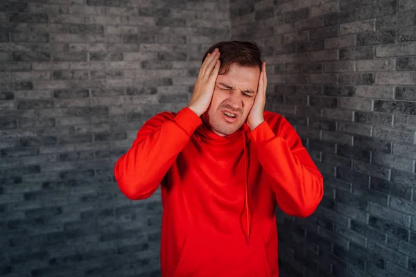 Un giovane in una felpa rossa afferra la testa con le mani in dolore su uno sfondo di mattoni. Un maschio ha mal di testa. — Foto Stock