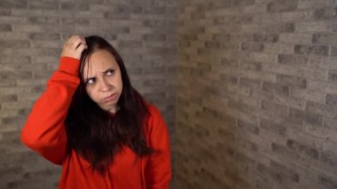 Kırmızı kapüşonlu güzel, duygusal bir kadın kafasını tuğla bir arka planda kaşıyor..