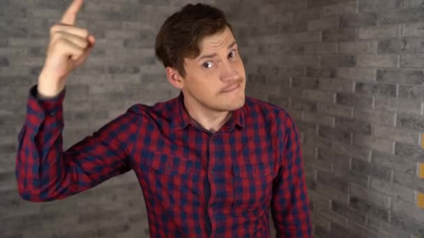 Ein junger gutaussehender Mann im karierten Hemd zeigt mit dem Finger in die Kamera auf grauem Backsteinhintergrund. — Stockvideo